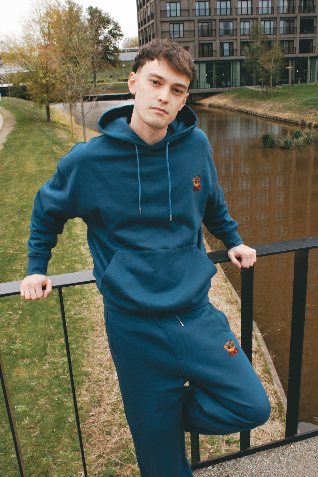 Person wearing a blue tracksuit with a logo, standing by a river with buildings in the background.